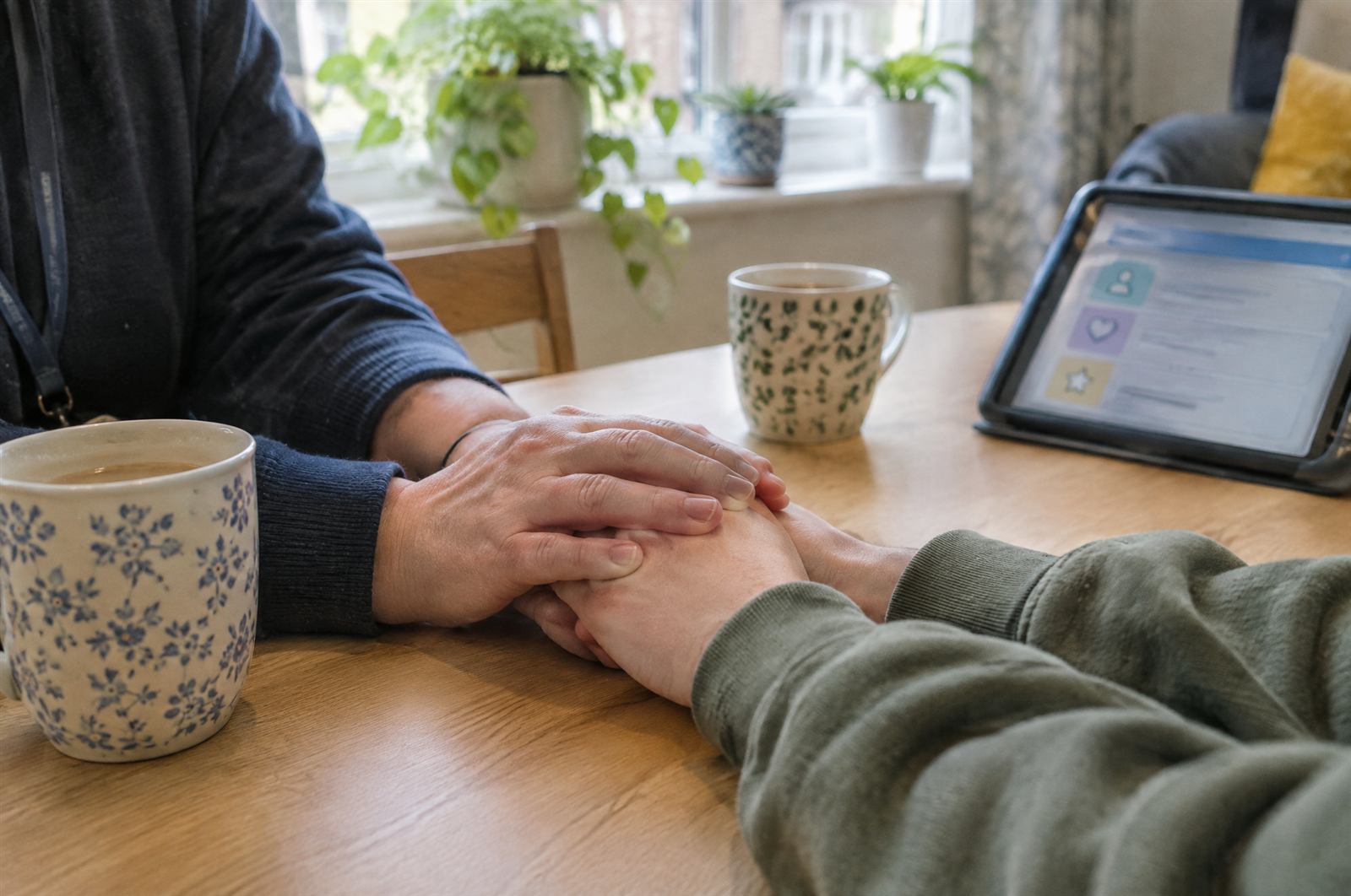 A support worker’s hands resting over a young person’s hands at a kitchen table, beside a tablet